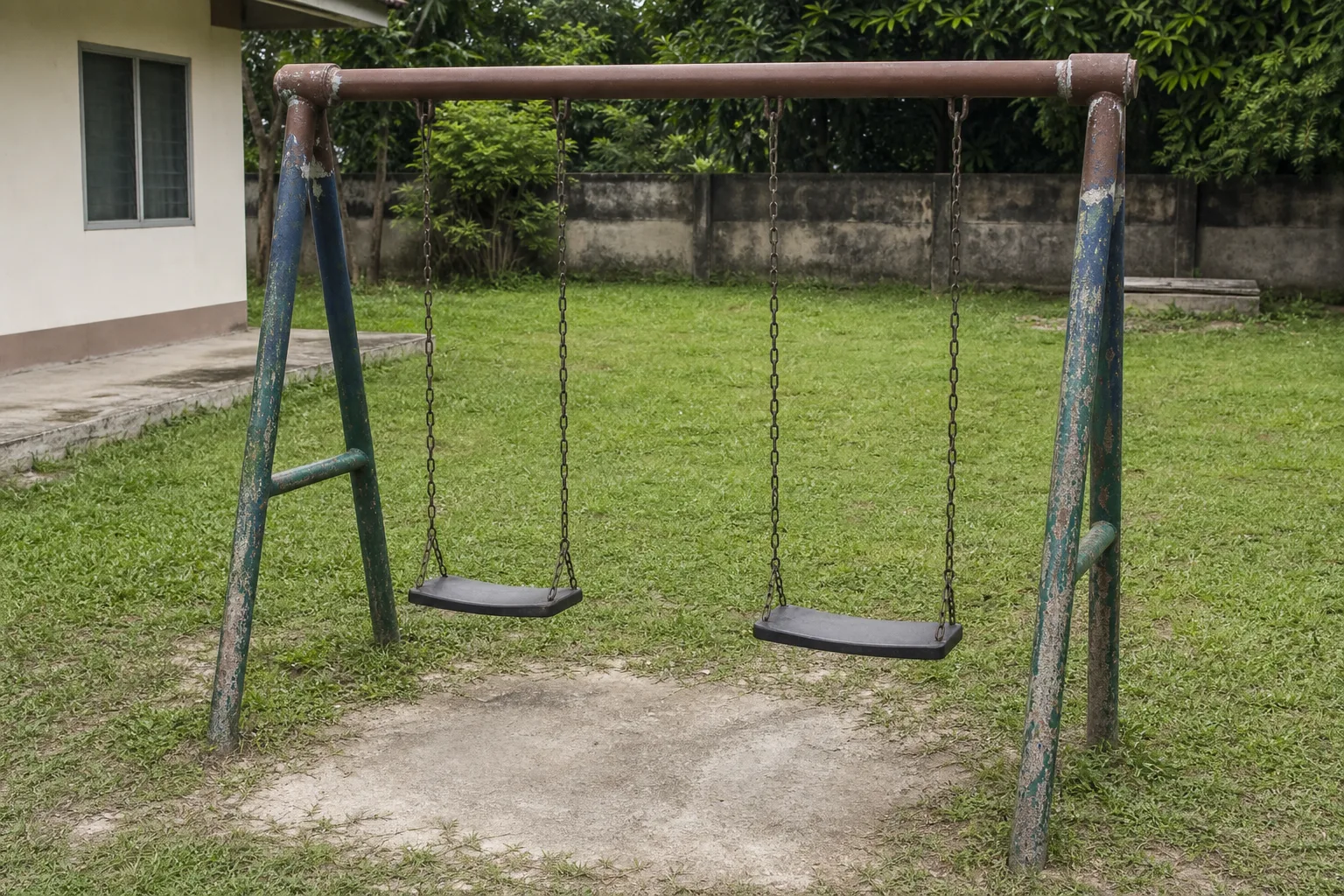 An old steel swing frame on grass, its top rail freshly painted against weathered uprights, two seats hanging empty on their chains, nobody in the frame